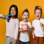 Three children are standing against a orange background. They are all smiling and holding up dental supplies to indicate they care about their teeth. There is one male and two females. The male is centered.