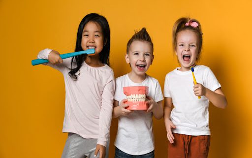 Three children are standing against a orange background. They are all smiling and holding up dental supplies to indicate they care about their teeth. There is one male and two females. The male is centered.