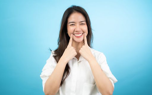 A woman is smiling with her hands pointing to her mouth. She looks happy and is wearing a white shirt.