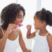 A mom and her daughter are practicing dental hygiene by brushing their teeth together and smiling.
