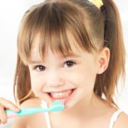 A young girl brushes her teeth as she smiles at the camera. She has brown hair done up in pigtails and brown eyes.